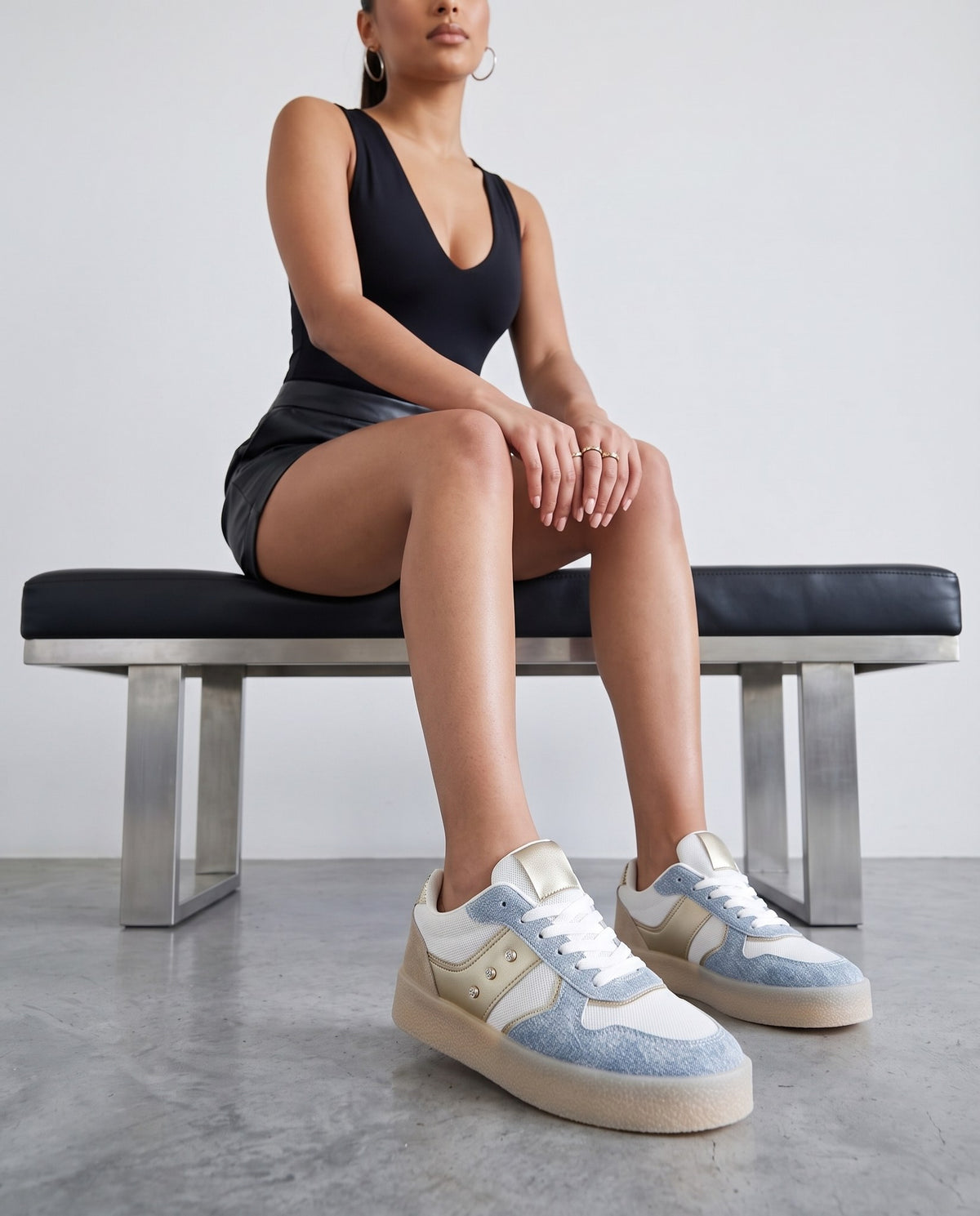 Modella seduta su panca nera, sneakers bianche e denim con dettagli oro in primo piano
