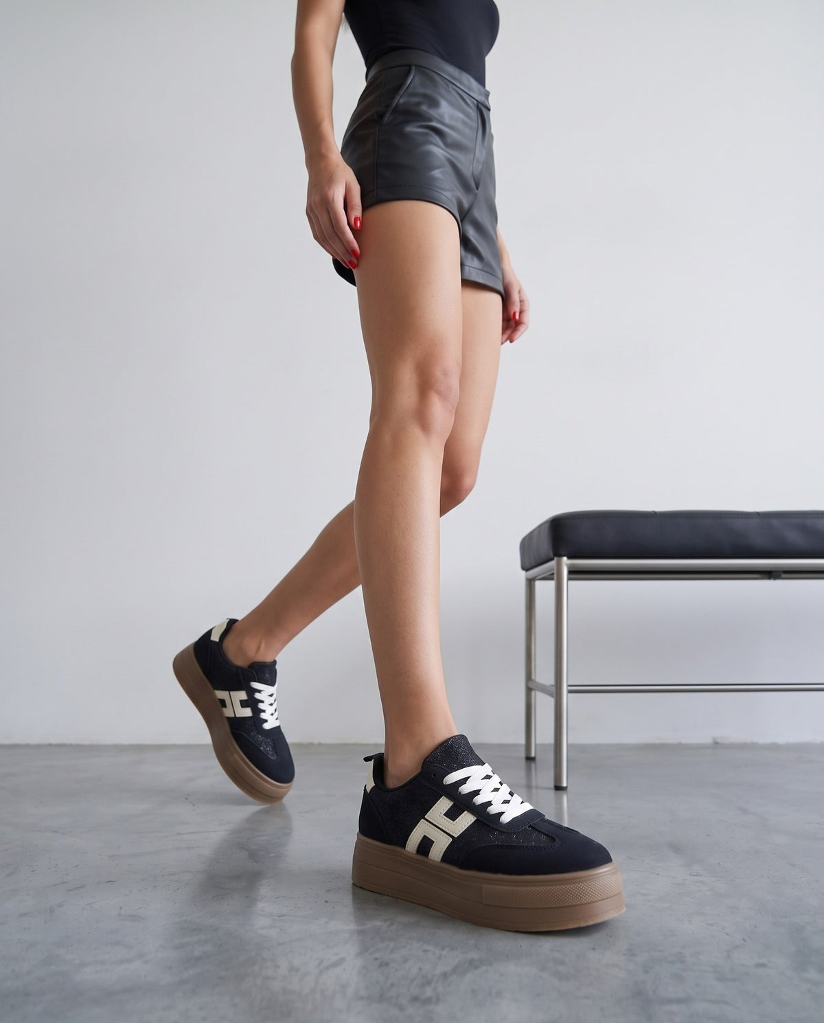 Gambe di modella con sneaker nere platform glitterate, logo bianco a H, suola gum, panca in acciaio e pelle sullo sfondo.