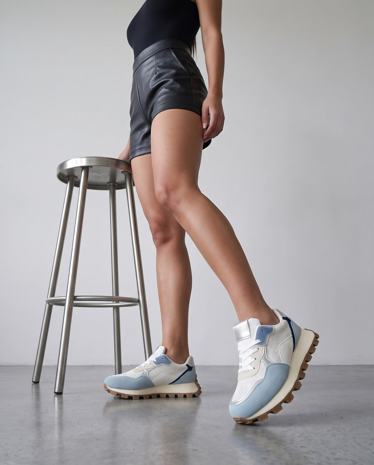 Gambe della modella con sneaker bianca e celeste, sgabello in acciaio, pavimento in cemento grigio chiaro.