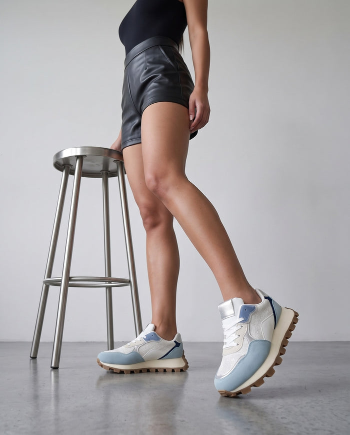 Gambe della modella con sneaker bianca e celeste, sgabello in acciaio, pavimento in cemento grigio chiaro.
