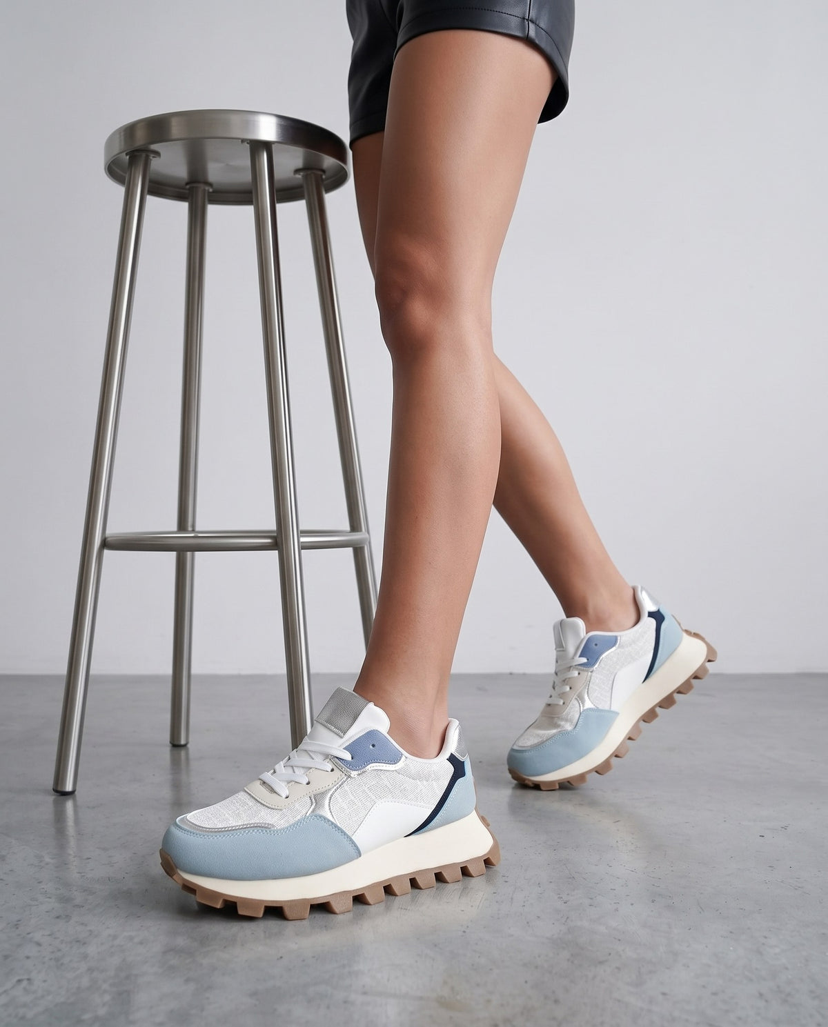 Dettaglio ravvicinato delle gambe e delle sneakers bianche con inserti celesti vicino a uno sgabello metallico
