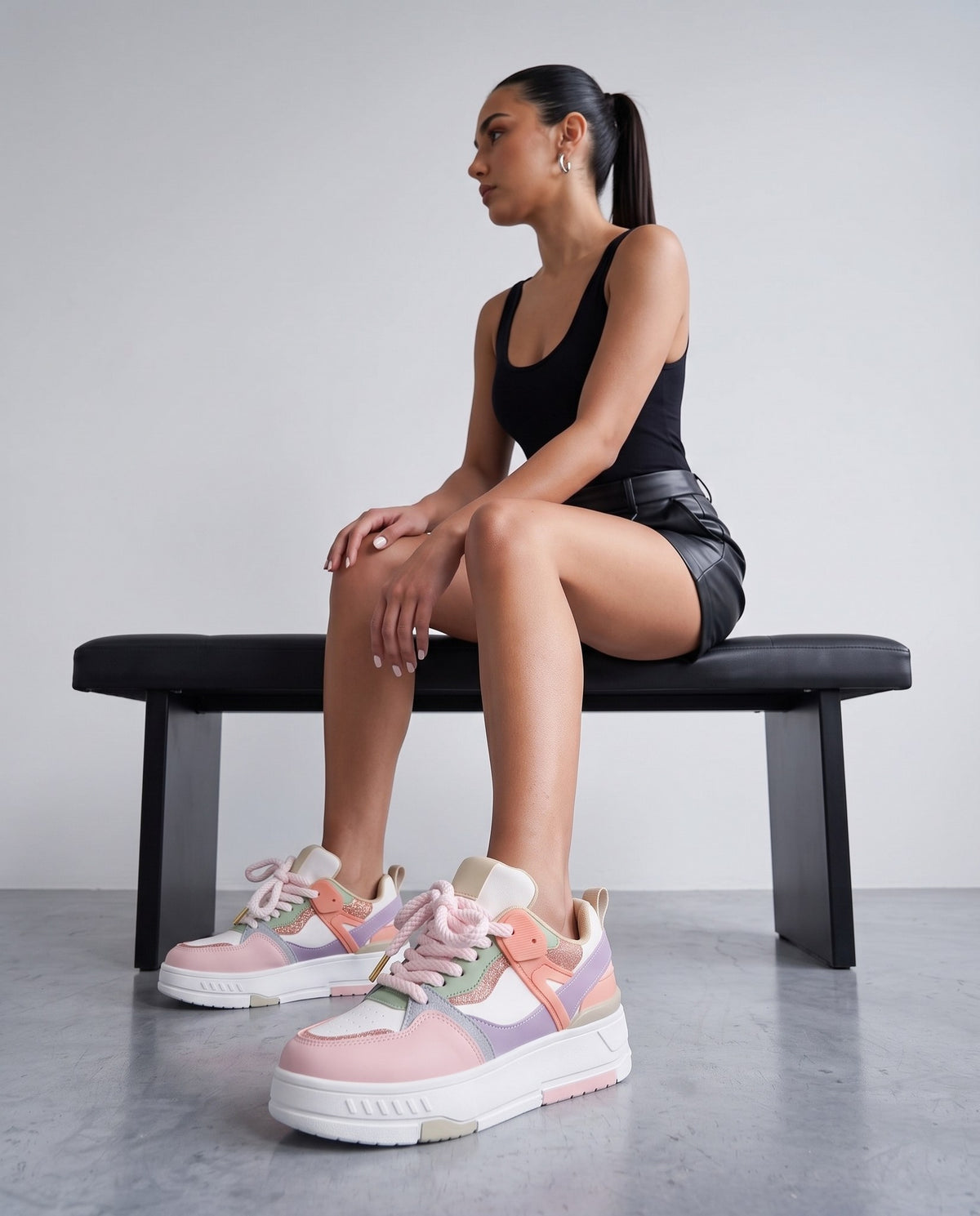 Modella seduta su panca nera, sneakers multicolor pastello in primo piano, sfondo grigio chiaro da studio.