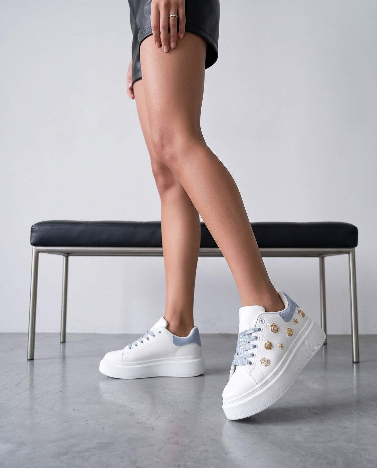 Dettaglio ravvicinato di sneaker bianche con charm dorati, lacci e tallone azzurri, su gambe in primo piano.