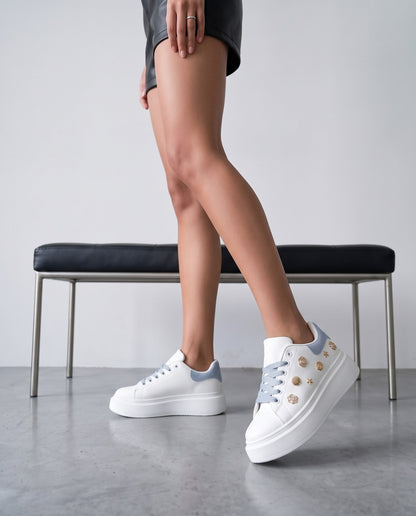 Dettaglio ravvicinato di sneaker bianche con charm dorati, lacci e tallone azzurri, su gambe in primo piano.