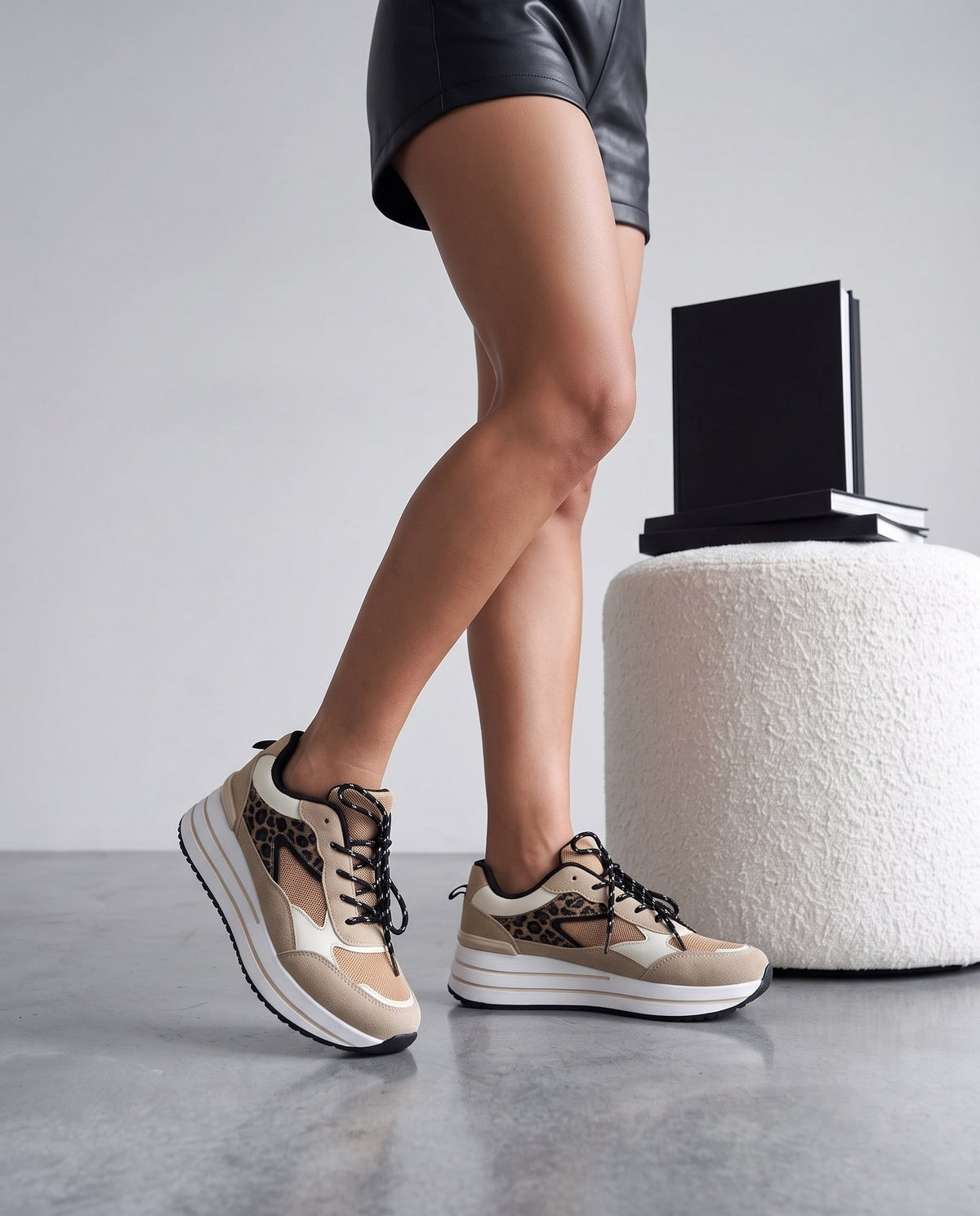 Gambe in piedi con sneaker leopardate beige, pouf bianco e libri neri sullo sfondo.
