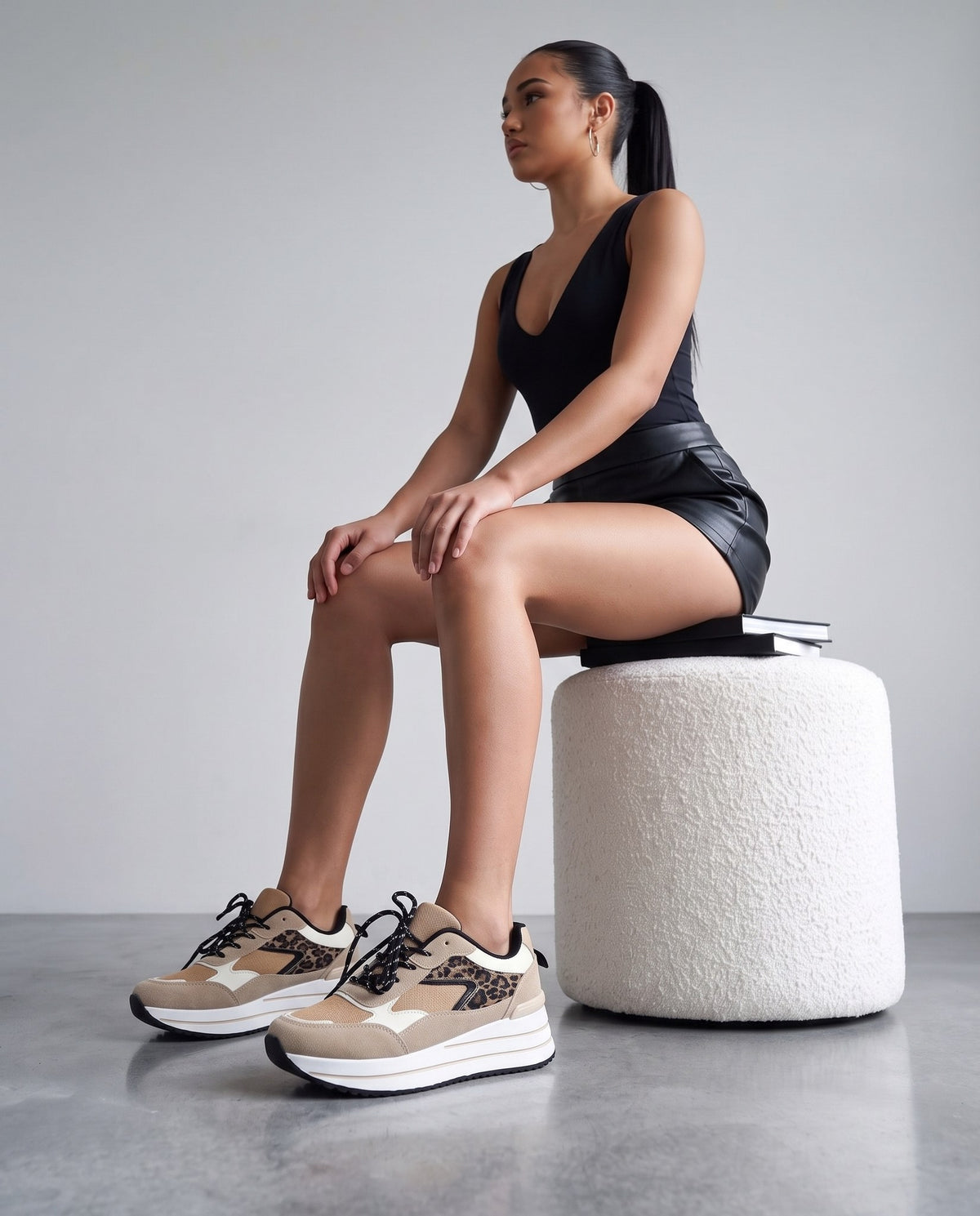 Modella seduta su pouf bianco, gambe accavallate, sneakers leopardate beige in primo piano.