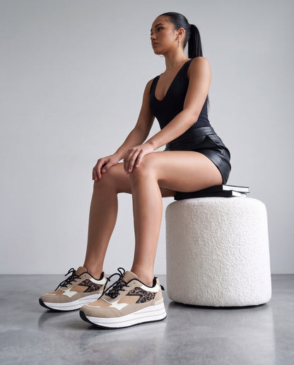 Modella seduta su pouf bianco, gambe accavallate, sneakers leopardate beige in primo piano.