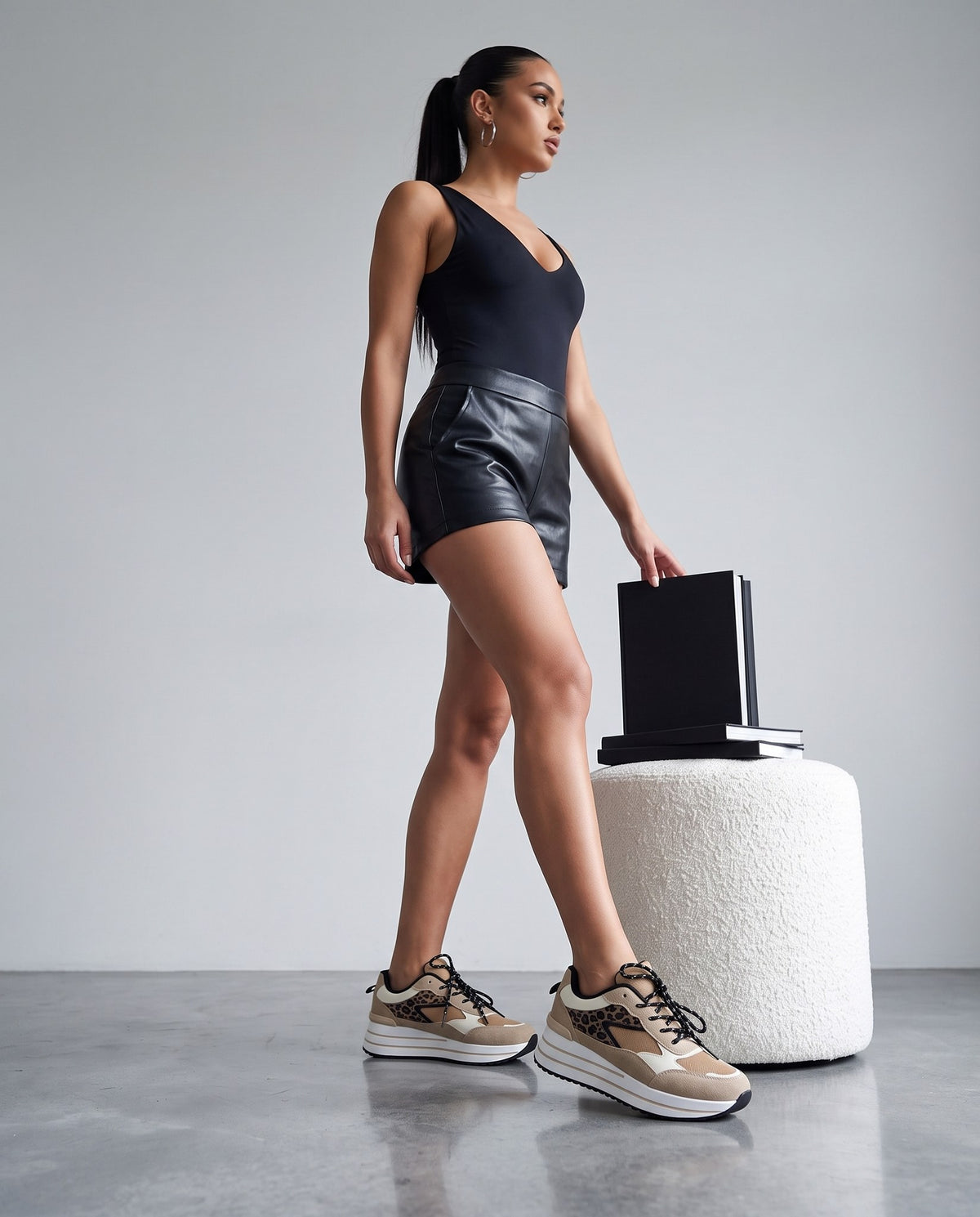 Modella in body nero e shorts in ecopelle con sneaker leopardate beige, accanto a pouf bianco con libri.