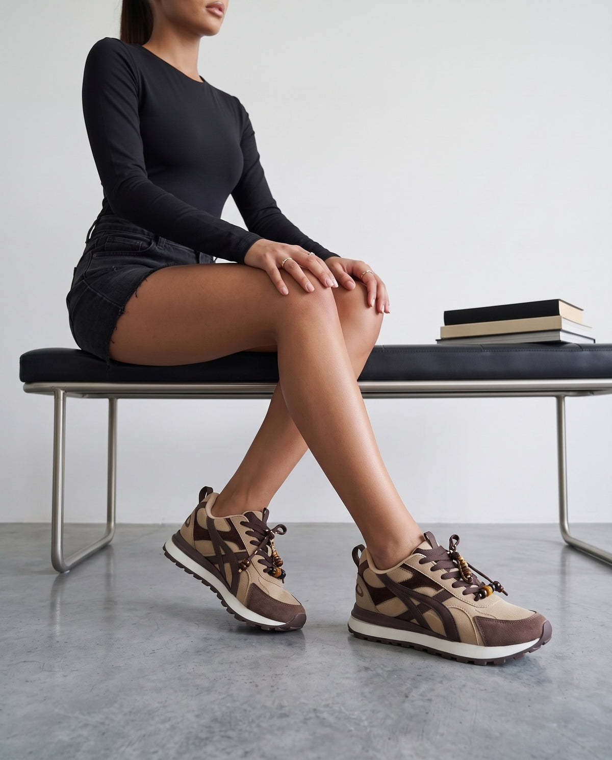 Modella seduta su panca nera, gambe accavallate, sneakers beige e marrone con charm di perline in primo piano.