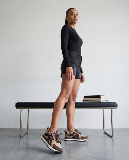 Modella in body nero e shorts denim cammina verso la camera con sneakers beige e marroni con charm, accanto a panca con libri.