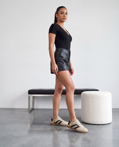 Modella in piedi di profilo con sneakers khaki glitter oro e inserto cow-print, body nero e shorts in ecopelle.