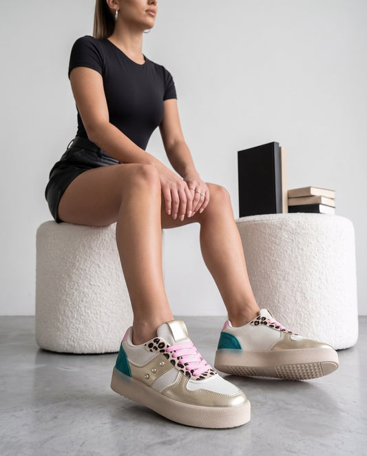 Modella seduta su pouf bianco, gambe distese, sneakers multicolor oro/bianco con dettagli leopardati in primo piano.
