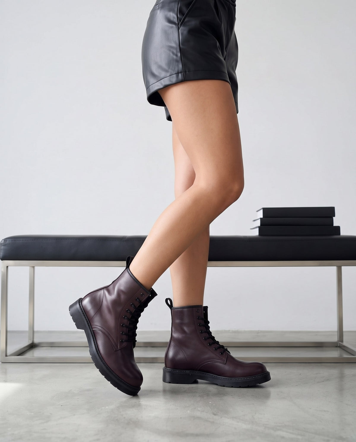 Gambe femminili con stivali bordeaux in pelle, panca con libri sullo sfondo, pavimento in cemento grigio.