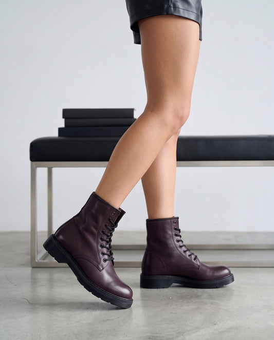 Dettaglio di due stivaletti bordeaux in pelle con lacci neri, gambe della modella in primo piano, panca sullo sfondo.