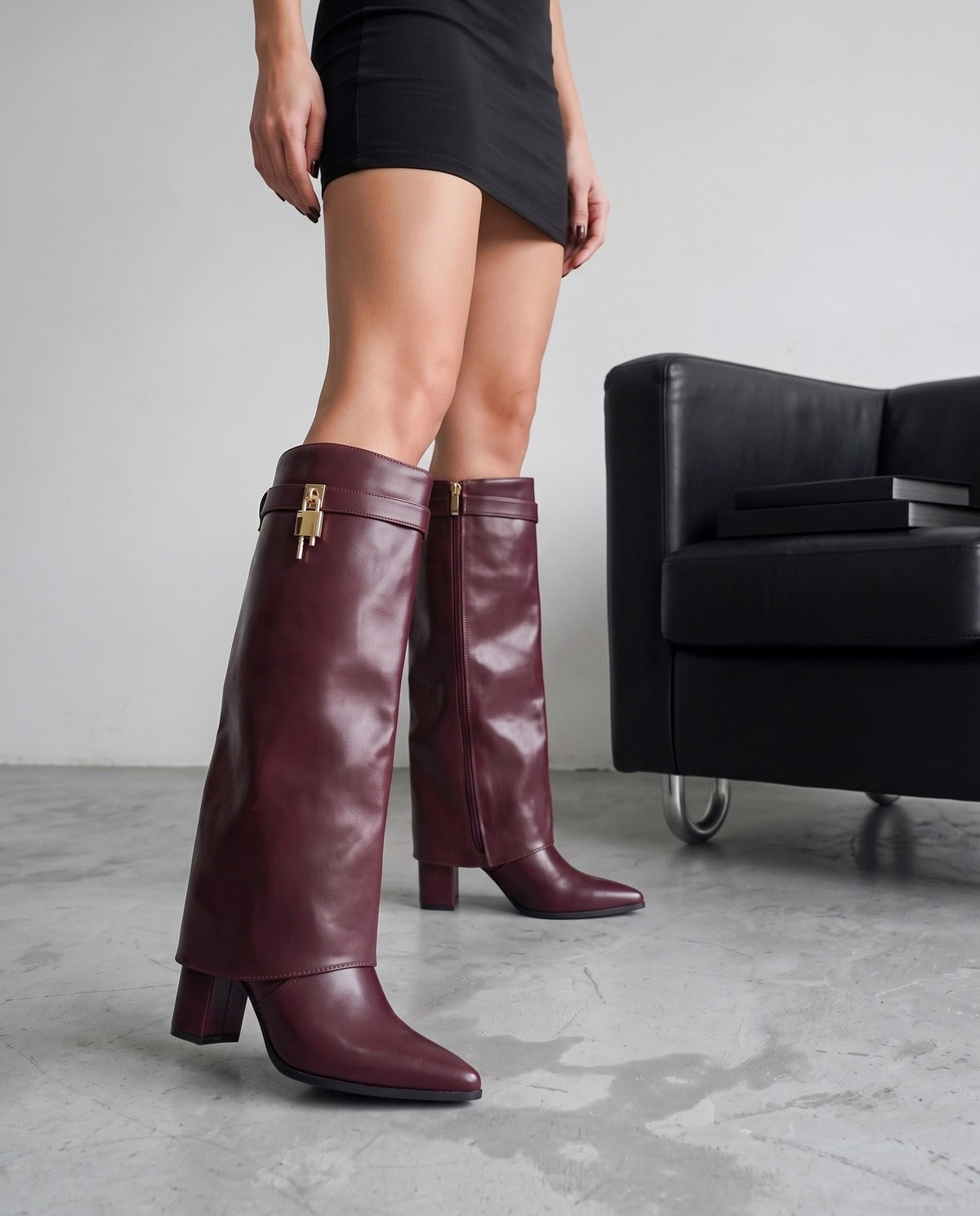 Gambe femminili con stivali bordeaux a risvolto, tacco alto, accanto a poltrona nera con libri