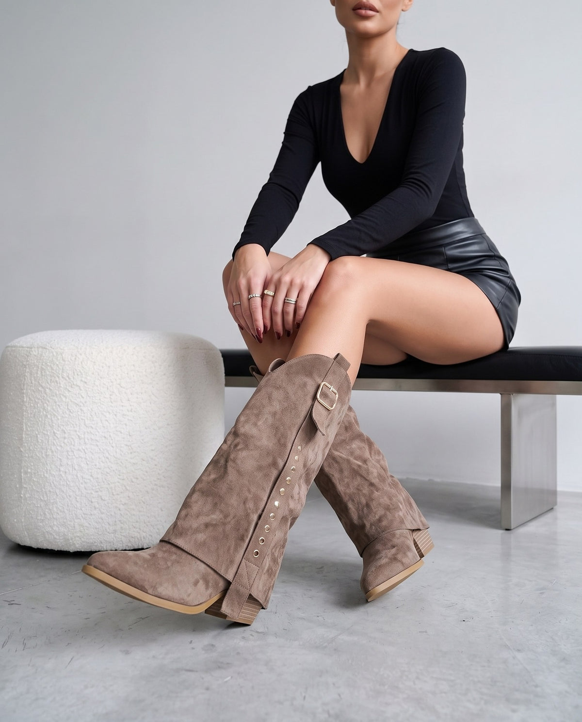 Modella seduta su panca, gambe accavallate, stivali scamosciati color tortora con fibbia e borchie in primo piano