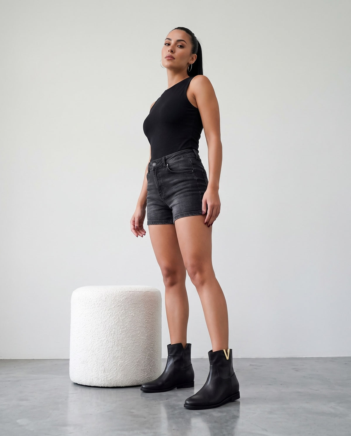 Modella in piedi con stivaletti neri con placca dorata a V, shorts di jeans e top nero, accanto a pouf bianco