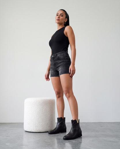 Modella in piedi con stivaletti neri con placca dorata a V, shorts di jeans e top nero, accanto a pouf bianco