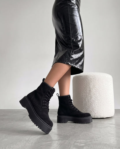 Gambe con stivaletti neri in camoscio, plateau carrarmato, accanto a pouf bianco bouclé su pavimento in cemento