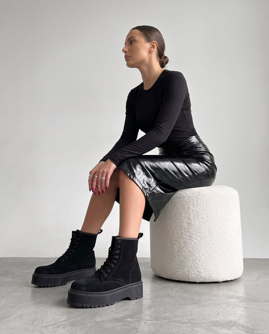 Modella seduta su pouf bianco, stivaletti in camoscio nero platform in primo piano