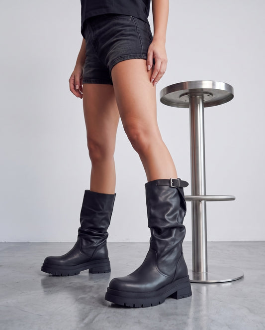 Gambe in piedi con stivali neri slouchy, accanto a uno sgabello cromato, su pavimento in cemento