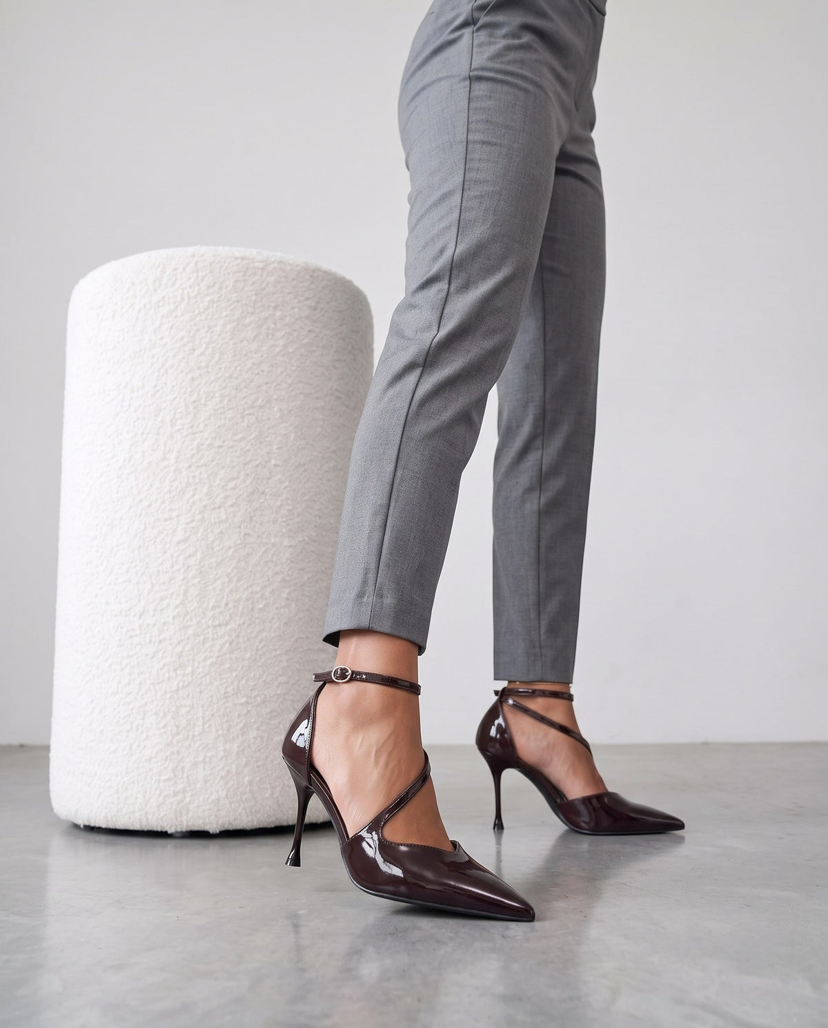 Gambe con pantaloni sartoriali grigi e décolleté bordeaux lucide accanto a pouf bianco bouclé