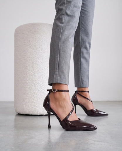 Primo piano di gambe con décolleté in vernice marrone scuro, cinturino alla caviglia, accanto a pouf bianco
