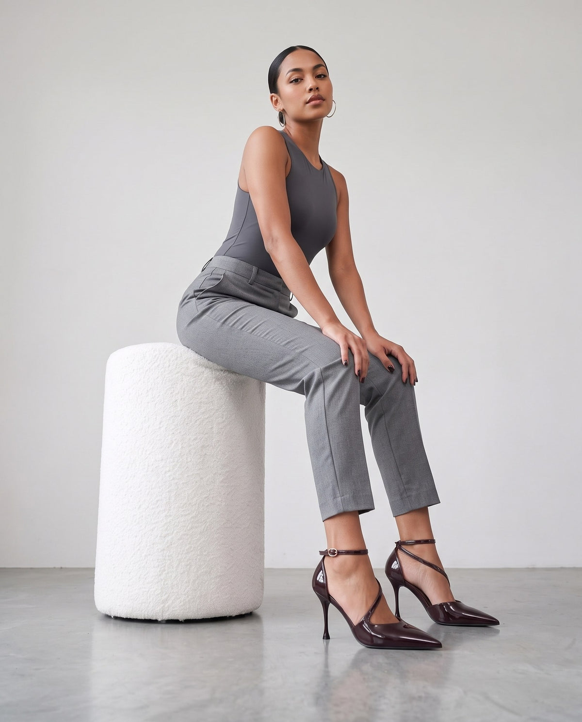 Modella seduta su pouf bouclé bianco, décolleté bordeaux scuro con cinturino alla caviglia, primo piano sulle scarpe