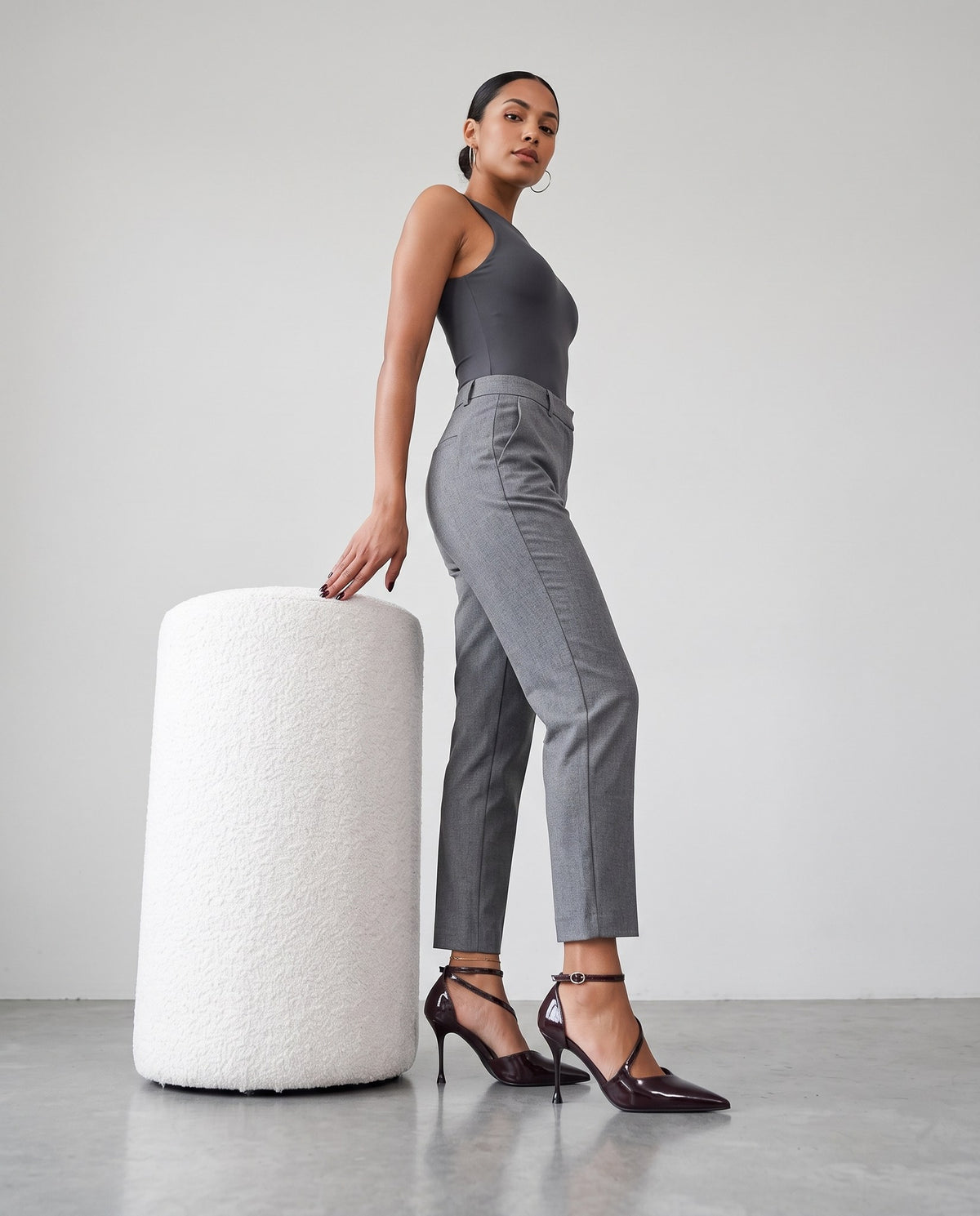 Modella in piedi con tacchi décolleté marroni lucidi, pantaloni grigi e body antracite, accanto a un pouf bianco.