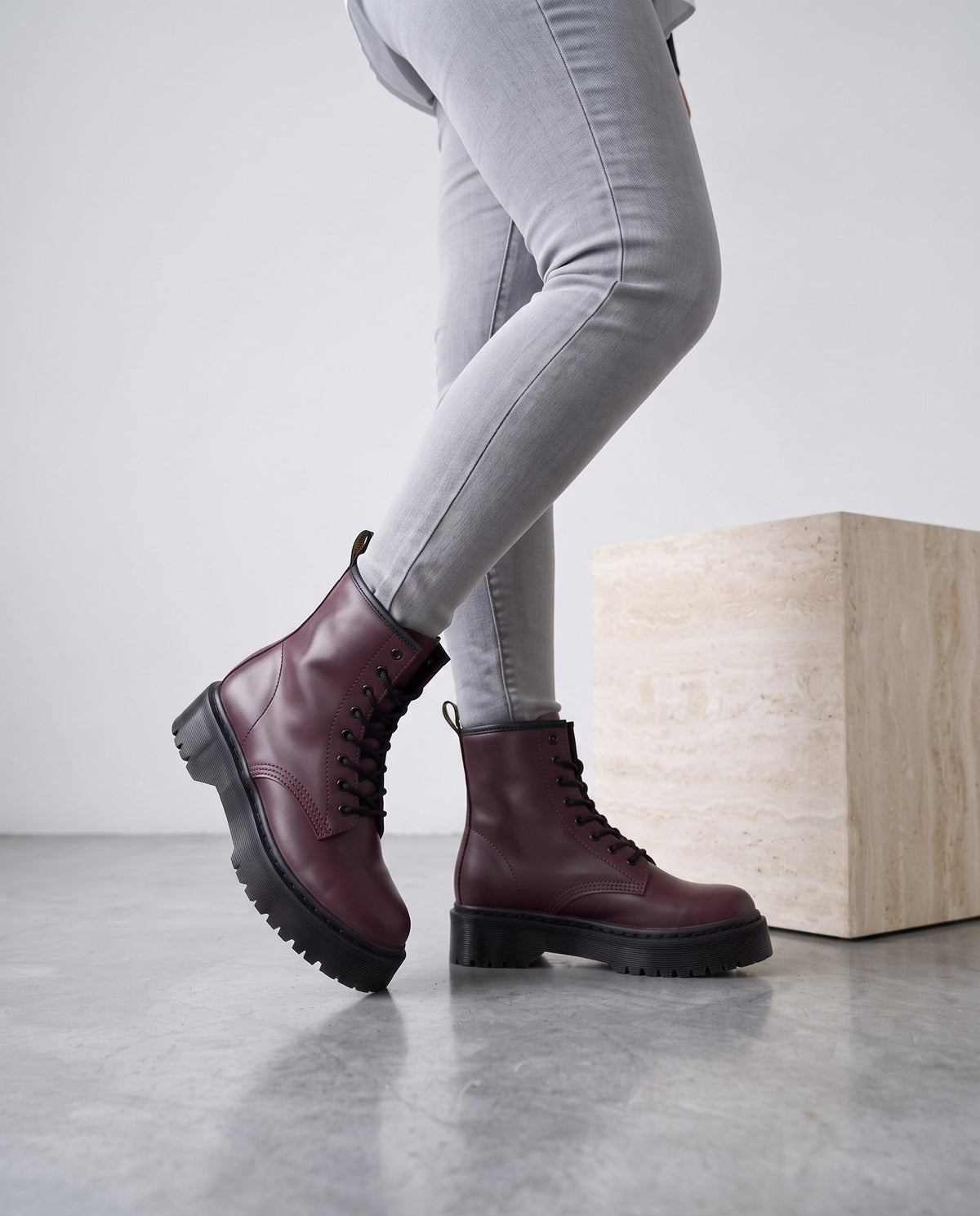 Gambe con jeans grigi e stivaletti bordeaux in posa, accanto a cubo di travertino su pavimento in cemento