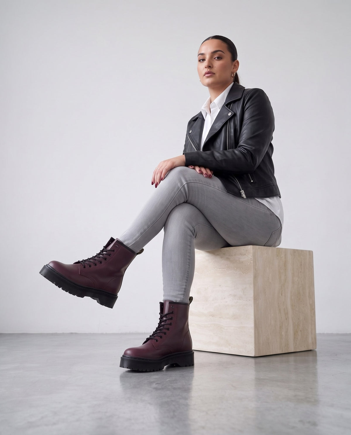 Modella seduta su cubo in travertino con stivaletti bordeaux stile combat, gambe accavallate, outfit grigio e giacca di pelle nera