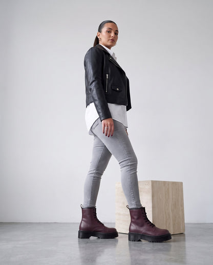 Modella di profilo in jeans grigi, giubbino di pelle nero e stivaletti bordeaux con suola carrarmato.