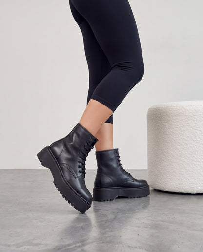 Gambe con stivali neri platform, pouf bianco bouclé a fianco, pavimento in cemento grigio