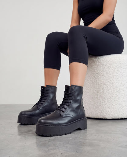 Modella seduta su pouf bianco, gambe accavallate, stivali platform neri in primo piano