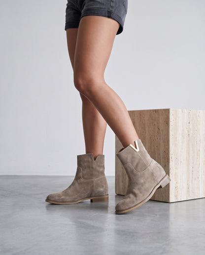 Gambe femminili con stivaletti in camoscio taupe, accanto a cubo in travertino, su pavimento in cemento