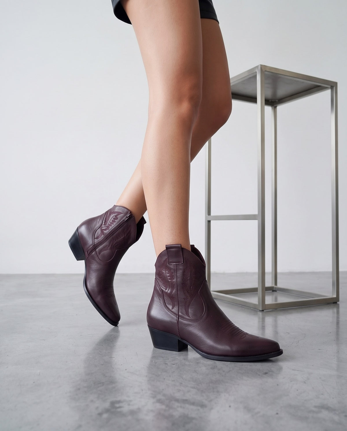 Gambe in posa di cammino con stivaletti texani bordeaux, tavolino in acciaio sullo sfondo