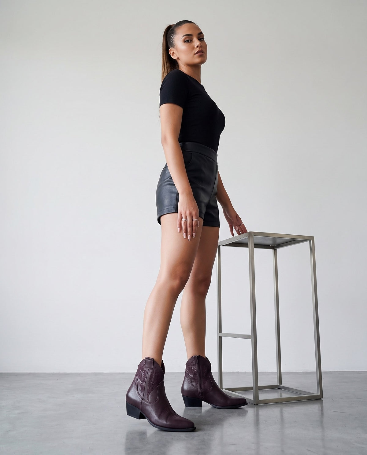 Modella di profilo con stivaletti texani bordeaux, shorts e top neri, accanto a sgabello in acciaio.