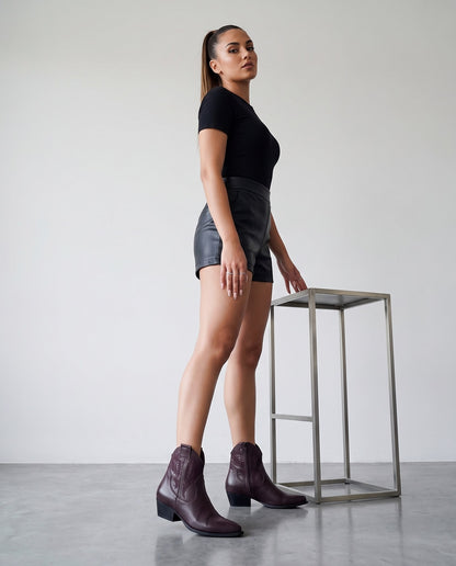 Modella di profilo con stivaletti texani bordeaux, shorts e top neri, accanto a sgabello in acciaio.