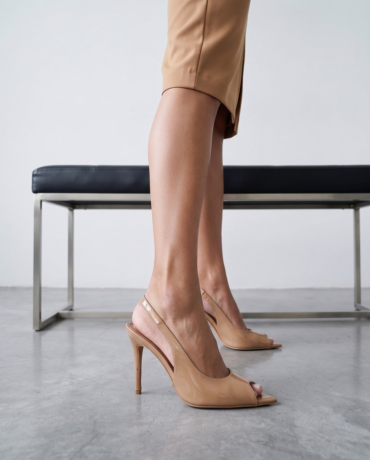 Gambe femminili con sandali slingback beige a punta aperta, tacco a stiletto, accanto a panca con telaio cromato.