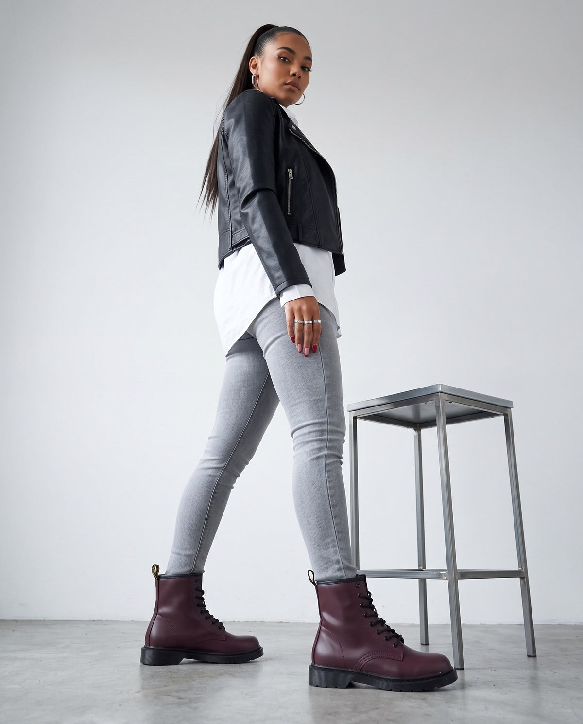 Modella di profilo in piedi con stivaletti bordeaux stile combat boot, accanto a uno sgabello in acciaio