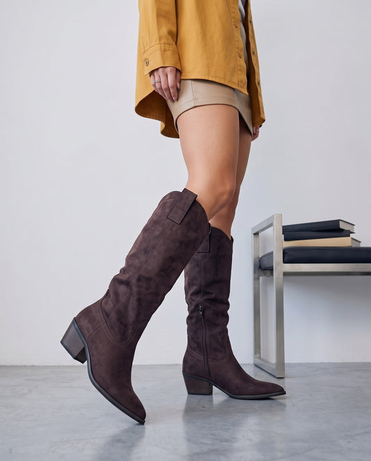Gambe con stivali camperos marrone scuro in camoscio, posa in cammino, panca con libri sullo sfondo