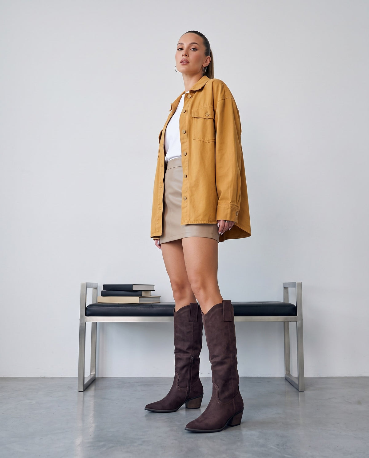 Modella in piedi con stivali camperos in camoscio marrone, outfit senape e minigonna beige, panca con libri sullo sfondo.