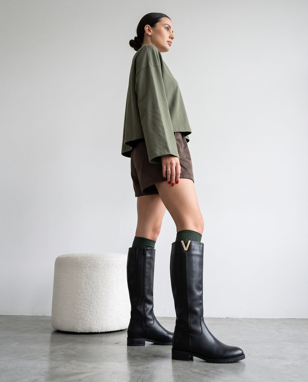 Modella di profilo con stivali neri al ginocchio, maglia verde oliva e shorts marroni, accanto a pouf bianco