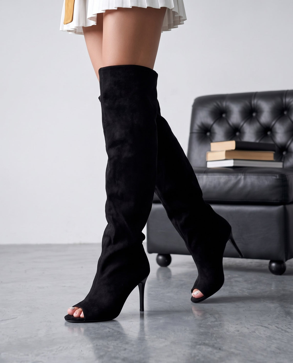 Primo piano di gambe con stivali cuissard neri in camoscio, poltrona nera sfocata sullo sfondo