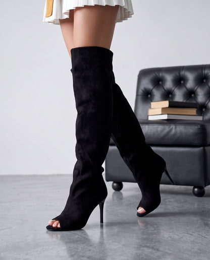 Primo piano di gambe con stivali cuissard neri in camoscio, poltrona nera sfocata sullo sfondo