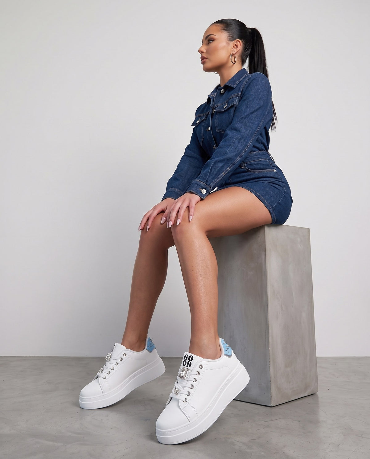 Modella seduta su blocco di cemento, sneakers bianche con dettaglio azzurro e gemme in primo piano