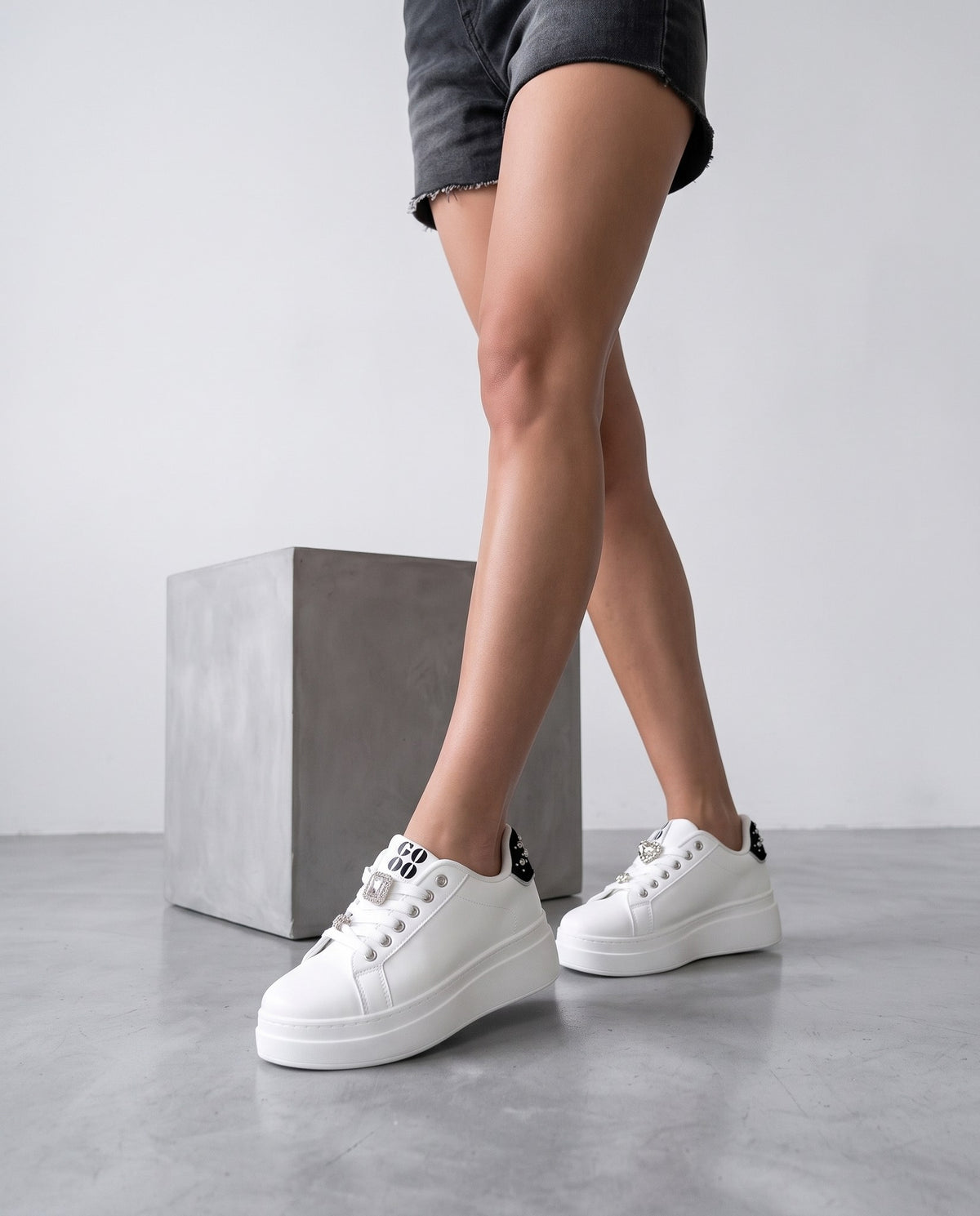 Gambe di modella in piedi con sneaker bianche platform, vicino a un blocco in cemento grigio