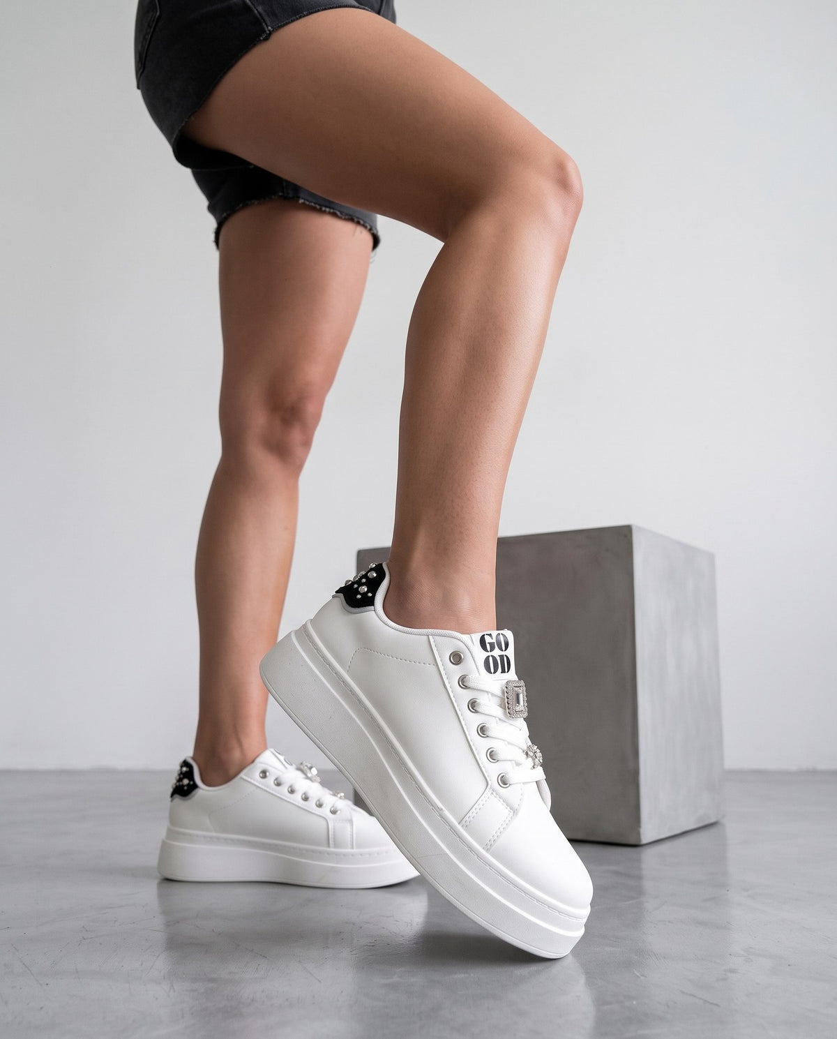 Primo piano scarpe sneaker bianche con dettagli gioiello, gambe della modella su sfondo grigio con cubo in cemento