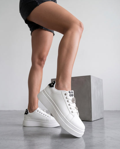 Primo piano scarpe sneaker bianche con dettagli gioiello, gambe della modella su sfondo grigio con cubo in cemento