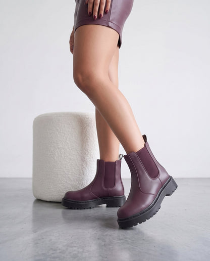 Gambe con stivaletti Chelsea bordeaux, pouf bianco bouclé sullo sfondo, pavimento in cemento grigio.